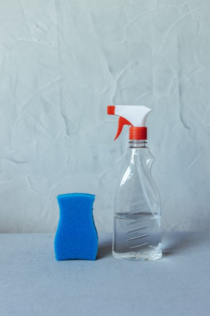 A clear plastic spray bottle filled with cleaning solution, featuring an orange and white trigger spray head, positioned next to a blue sponge on a light grey surface. The background is a textured, li
