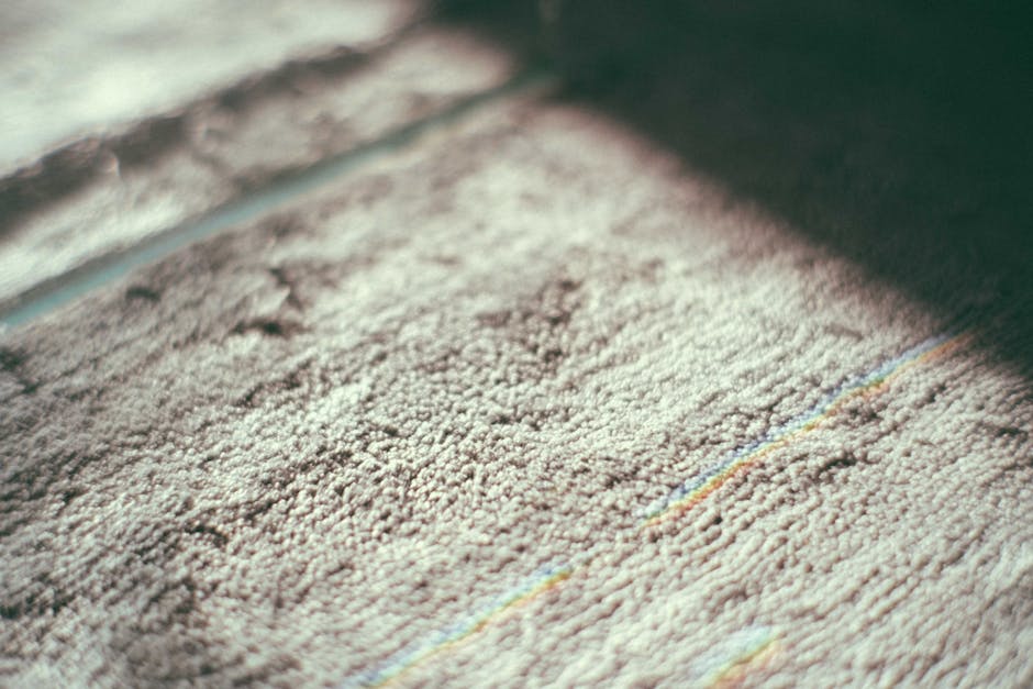 A woman with curly hair, dressed in a beige jacket, blue jeans, and white sneakers, is engaged in surface cleaning using a yellow vacuum cleaner with a black hose attachment on a patterned area rug in
